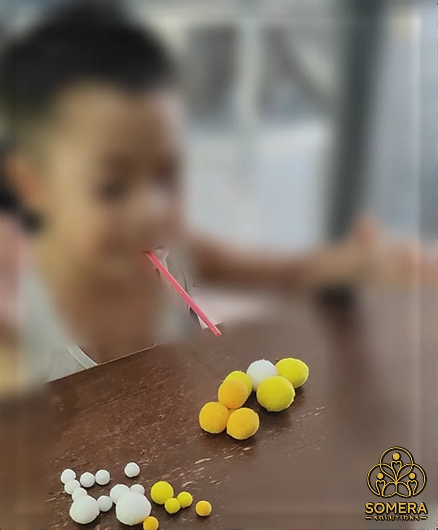 Child blowing through a red straw to move yellow and white soft pom-pom balls across a wooden table, with the face and background blurred