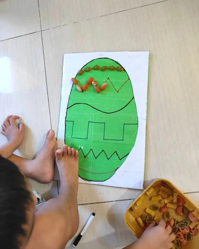 A child's hand placing colored pasta in a pattern on a green cardboard egg on the floor at home.