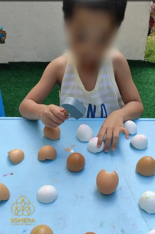 A child taps colored eggshells with a small hammer at a blue table